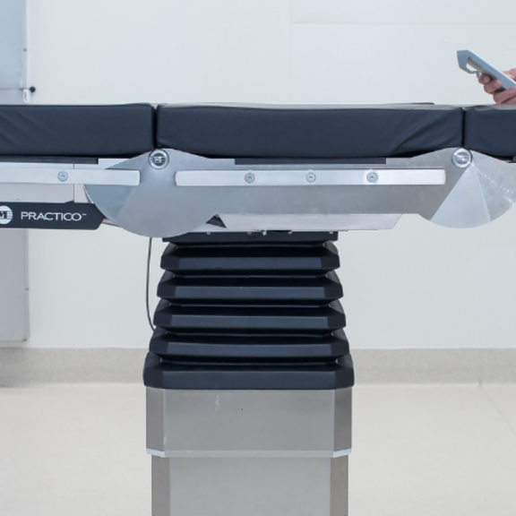 Silver and black operating table with a white background and a surgeon wearing a blue gown using the table 
