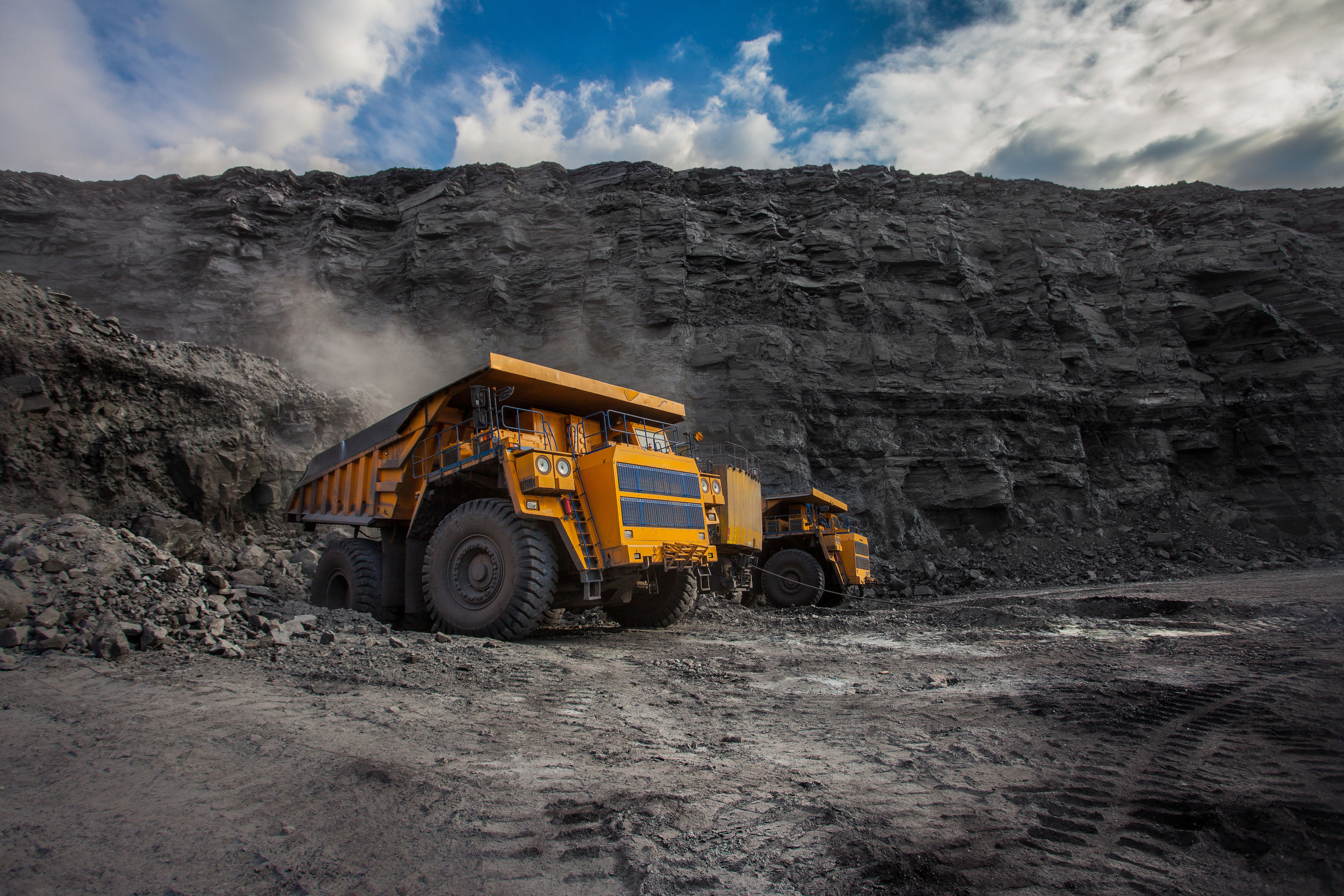 Mining site with a large yellow vehicle transporting materials, surrounded by rugged terrain and other equipment.