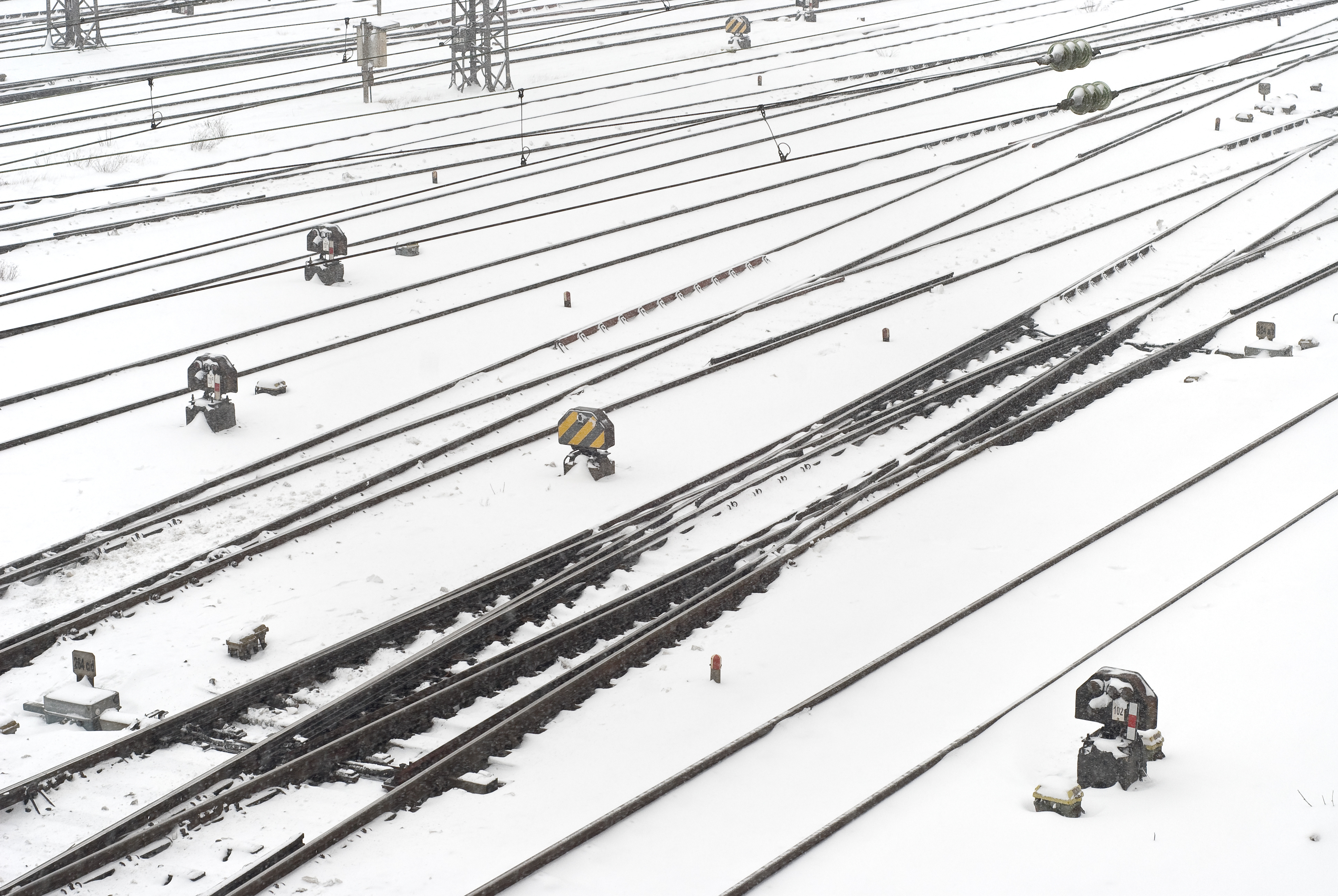 An image of a railway track featuring the points drive mechanism, partially covered in snow. Snow has settled on the surrounding gravel and sleepers, creating a contrast between the white frost and the dark steel of the tracks.