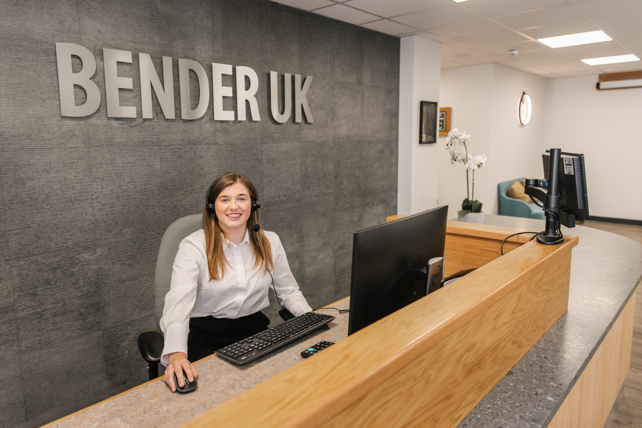 Image of receptionist sat on the reception desk with a desktop and laptop with a bender uk sign in the background